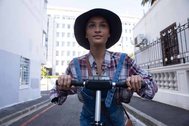Biracial alternative woman with short blonde hair, hat and denim dungarees out and about in the city on a sunny day, riding electric scooter. Urban independent woman on the go.