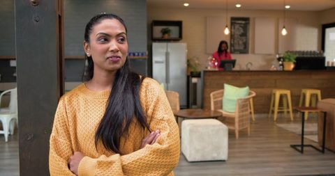 Indian woman leaning on wooden post in cozy cafe wearing mustard sweater, barista behind counter