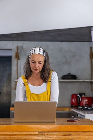 Middle-aged woman working remotely on laptop in rustic kitchen wearing yellow overalls