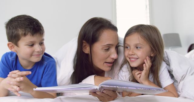 A mother is reading a storybook to her two children while lying in bed. They appear to be engaged and smiling, creating a warm and loving family moment. This image can be used to illustrate concepts of family bonding, reading and education, bedtime routines, and the joy of learning.