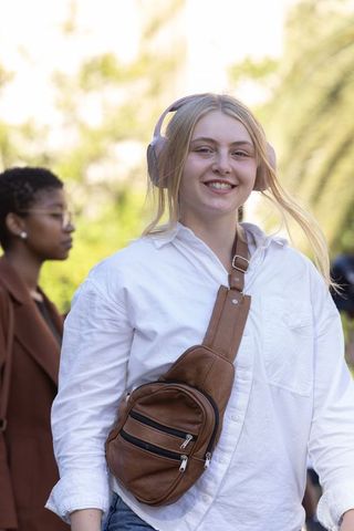 Blonde woman smiling while walking wearing pink headphones and leather sling bag on city street