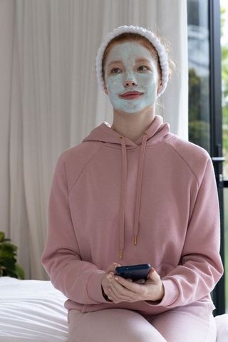 Young woman relaxing with mint face mask and smartphone in pink hoodie and headband