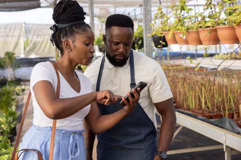 African american couple consulting with smartphone in greenhouse plant nursery