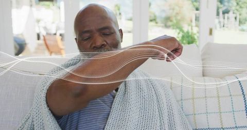 Senior man covering cough with elbow on sunlit sofa wrapped in knit blanket for home comfort