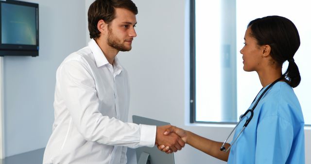A Caucasian man in business attire is shaking hands with an African American female nurse in a clinical setting, with copy space. Their handshake suggests a professional agreement or a gesture of thanks within a healthcare context.