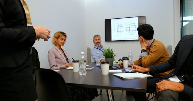 Group of business professionals sitting around table engaging in team discussion while one person presents data on screen. Ideal for use in business-focused content such as articles on collaboration, team meetings, project management strategies, and modern workplace environments.