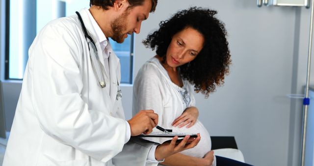 A Caucasian male doctor is consulting with a pregnant African American woman in a medical office, with copy space. They are reviewing information on a clipboard, discussing prenatal health or upcoming tests.