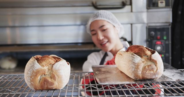 Woman baker in commercial kitchen smiling while taking freshly baked bread loaf from oven with tray. Image can be used in advertisements for bakeries, culinary schools, baking products, and food-related businesses. Great for illustrating fresh baked goods, catering services, and the baking profession.