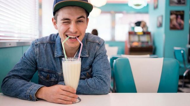 Young man sipping milkshake and winking in retro turquoise diner wearing denim jacket