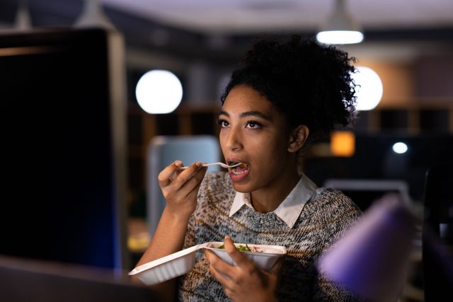 Young woman eating salad from a take-away box while working late in a creative office. Ideal for illustrating concepts of work-life balance, late-night work, office culture, healthy eating, and multitasking professionals.
