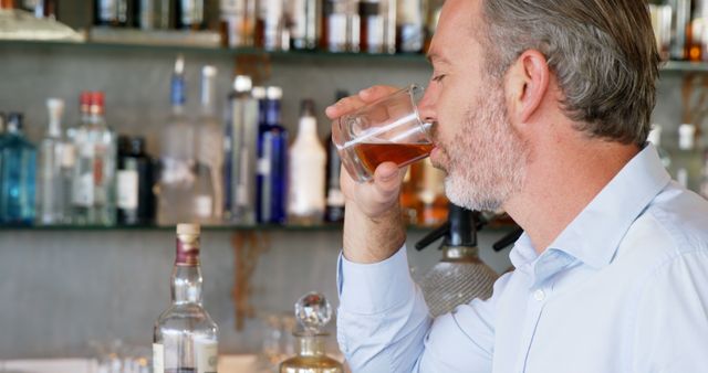 Mature man sipping whisky at a stylish bar with a variety of liquor bottles displayed in background. Ideal for use in lifestyle, nightlife, and hospitality industry content, promoting fine drinks and social settings, or illustrating concepts of leisure and relaxation.