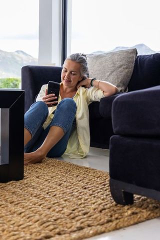Middle-aged woman sitting on jute rug using smartphone in bright contemporary living room