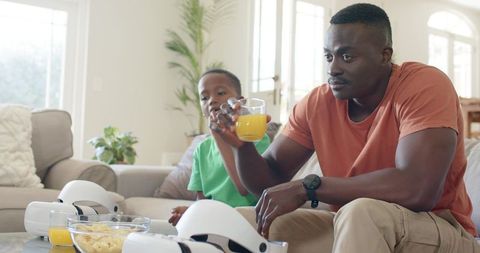 Father and Son Sharing Orange Juice and VR Headsets While Relaxing on Living Room Sofa