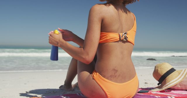 Rear view of biracial woman in bikini using sun lotion sitting on sunny beach by the sea - Download Free Stock Photos Pikwizard.com