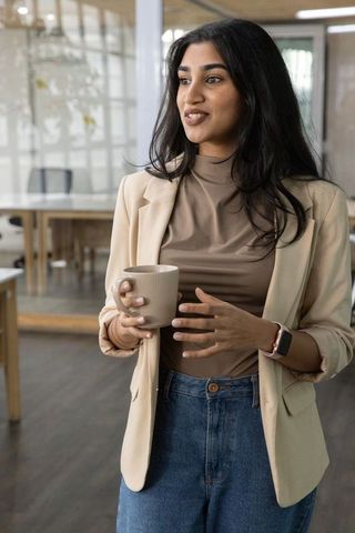 Indian professional woman holding mug wearing beige blazer and smartwatch in sunlit coworking