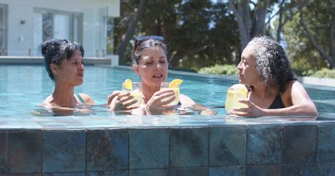 Three mature women enjoying cocktails and conversation in modern tiled pool at home