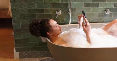 African american woman relaxing in bubble bath while using smartphone in modern bathroom