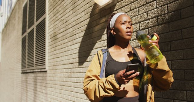 African american woman exercising outdoors drinking water and using smartphone in the city. healthy outdoor lifestyle fitness training.