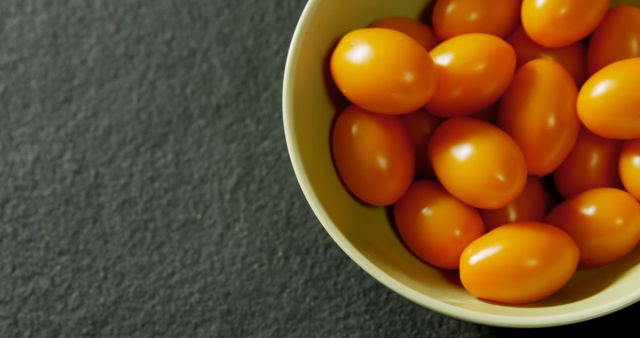A bowl filled with vibrant orange cherry tomatoes sits on a dark textured surface, with copy space - Download Free Stock Photos Pikwizard.com