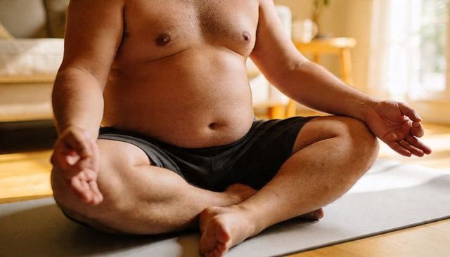 Plus-size man meditating shirtless on yoga mat in sunlit living room