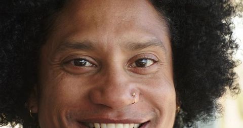 Smiling man with curly hair, gold nose ring and hoop earrings closeup headshot, warm smile