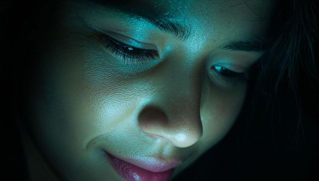 Woman gazing downward at smartphone teal glow closeup portrait low-key moody skin texture