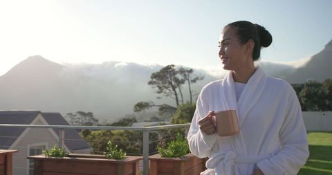 Mid-adult asian woman wearing white bathrobe enjoying sunrise on balcony holding ceramic mug