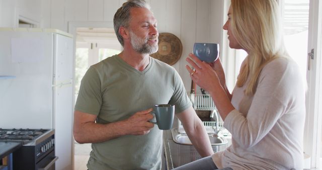 A mature couple happily drinking coffee in their home kitchen while having a morning conversation. The man is standing and holding a mug, while the woman is seated on the counter with a mug. Perfect for use in advertisements and articles about relationships, morning routines, and lifestyle content for mature audiences.