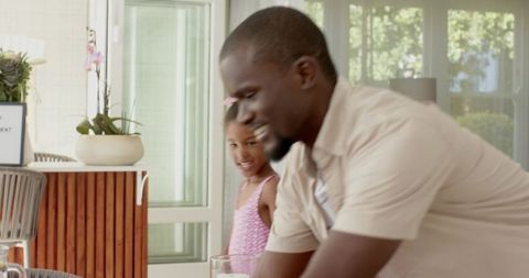 Father and daughter leaning over dining table placing glasses smiling in sunny sunroom