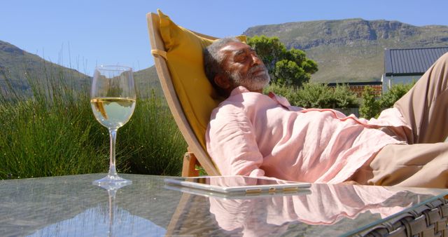 Senior man enjoying a sunny day outdoors, sleeping on a lounge chair with a glass of white wine nearby. Mountains and lush green landscape provide a serene background. Ideal for promoting relaxation, retirement lifestyles, peaceful living, outdoor leisure activities, and vacation homes.
