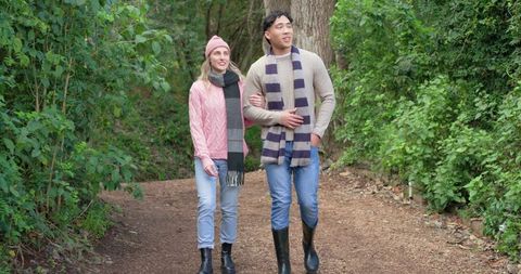 Couple strolling arm-in-arm on autumn woodland trail wearing cozy scarves, jackets and boots