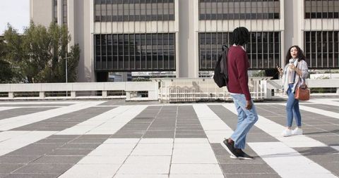 Diverse friends walking across urban plaza with striped tiles holding phone and coffee