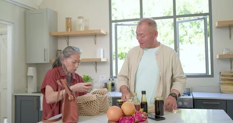 Senior asian couple unpacking groceries at sunlit kitchen island with reusable mesh bag