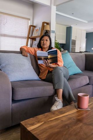 Mature asian woman reading paperback while sitting on gray sofa in cozy modern living room