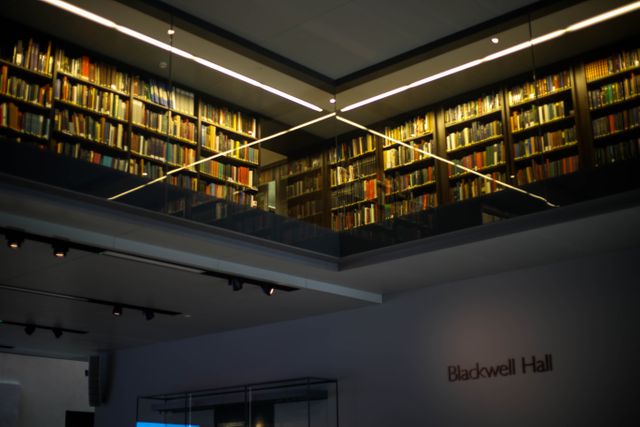 Interior view of a modern library with tall, floor-to-ceiling bookshelves filled with books. The sophisticated lighting focuses on the books, creating a warm and inviting atmosphere. Design features include sleek, minimalistic lines and glass barriers. Perfect for illustrating topics related to education, reading culture, libraries, and modern architecture.