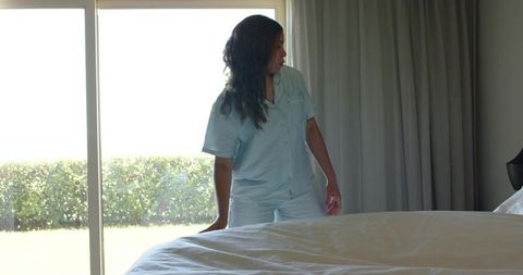 Woman in light blue pajamas standing by sliding patio door resting hand on bed
