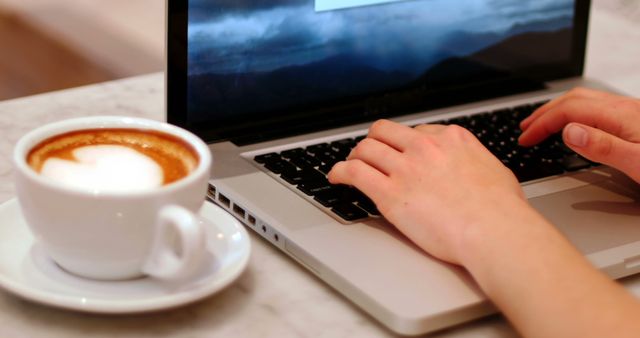 Hands seen typing on a laptop with a latte beside them on a table, suggesting a work or study routine in a coffee shop. Ideal for depicting freelancing, remote work, productivity, home office setups, coffee lovers.