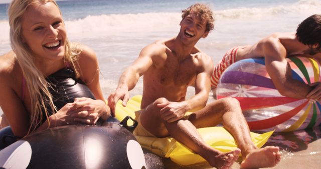 A group of young, Caucasian adults are enjoying a sunny day at the beach, laughing and sitting with inflatable rings. Their joyful expressions and beach attire capture the essence of a carefree summer outing with friends.