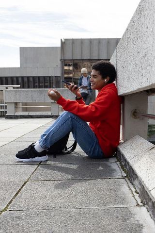 Urban student relaxing on rooftop terrace using smartphone and earbuds wearing red hoodie