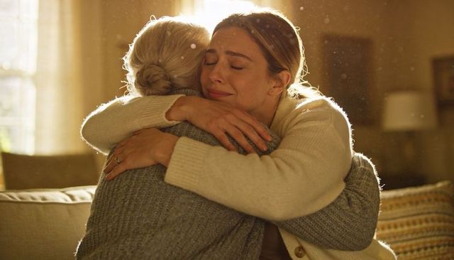 Adult daughter crying while embracing senior mother on cozy warm sunlit living room couch