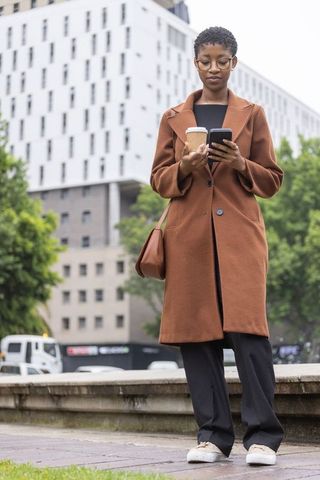 African professional woman standing in urban plaza checking phone and holding coffee