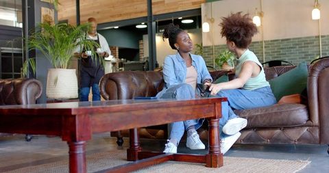Two African Women Sitting Chatting on Sofa in Cozy Relaxed Cafe Lounge with Greenery