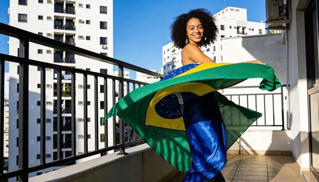 Spinning hispanic woman on sunlit balcony holding brazilian flag, celebrating brazil
