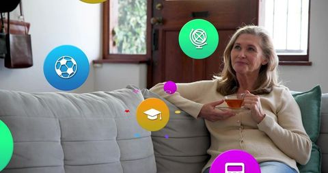 Senior woman relaxing with tea on sofa in living room with colorful floating app icons