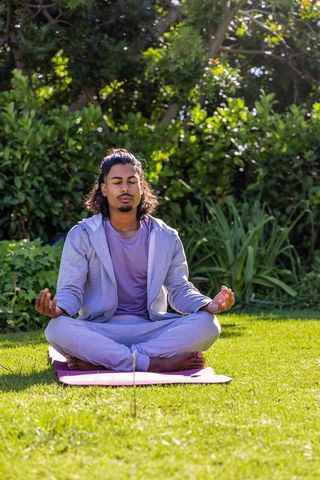 African american man meditating outdoors on pink yoga mat in sunlit garden, wearing hoodie