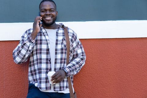 African american man smiling, talking on smartphone holding coffee while leaning on two-tone wall