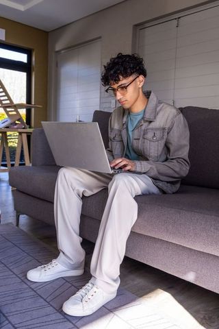 Young man working on laptop on grey sofa in modern living room wearing glasses