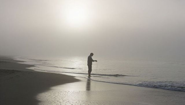 Fisherman standing in foggy dawn on empty beach with reflective wet sand and solitude