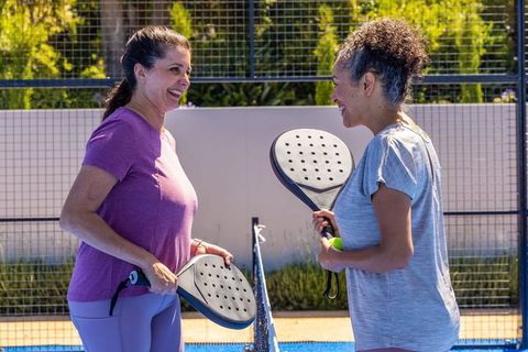 Diverse women smiling and chatting on outdoor padel court holding rackets and ball