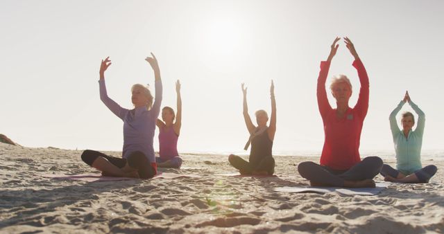 Senior women practicing yoga on sandy beach during sunrise. They are sitting in lotus position with arms raised, enjoying morning exercise. Perfect for promoting healthy lifestyle, senior fitness programs, and outdoor wellness activities.